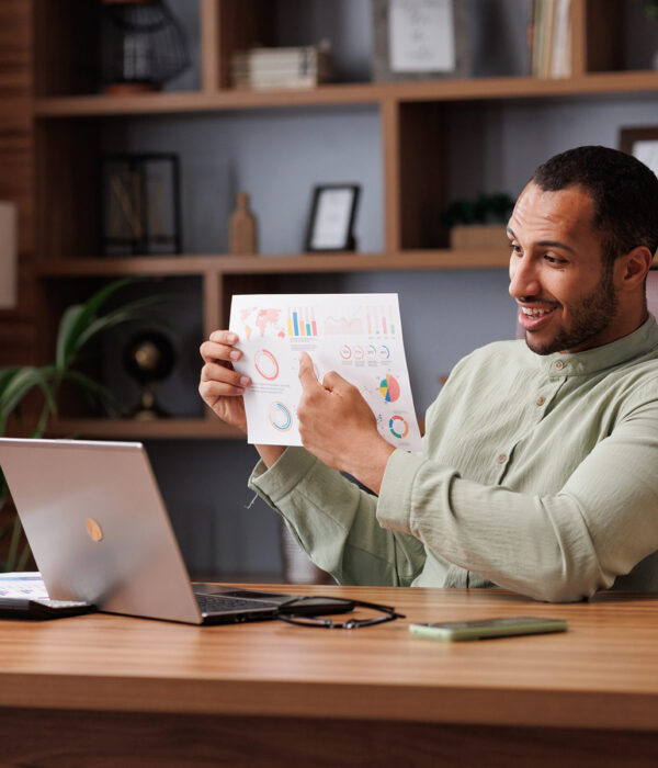 Young smiling mixed race businessman have video call sitting in office, showing financial report with graphs and charts to colleagues, partners and investors. Online video conference.