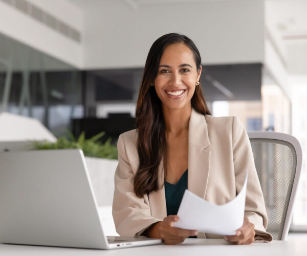 Portrait of happy female accountant or secretary sitting at desk with laptop, holding documents, making analysis, prepare agreement, review contract details, feel satisfied with productive workflow