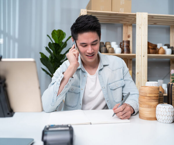 Asian young man talk with customer on phone to check vase goods order. Attractive business male work to prepare parcel boxes, checking ecommerce shipping online retail to sell at home warehouse store.